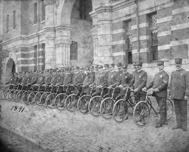 Two rows of uniformed police officers with bicycles lined up in front of the headquarters, 1899.